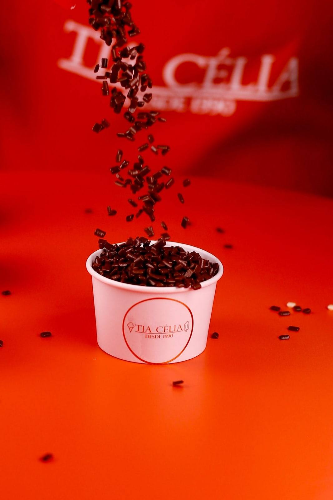 Granulado de chocolate caindo sobre um potinho de brigadeiro de colher da Tia Célia, em fundo vermelho.