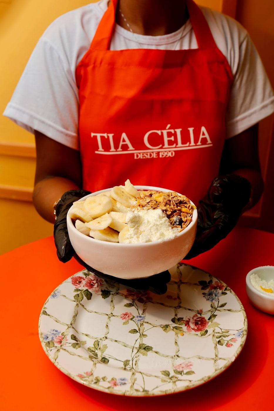 Atendente da Tia Célia de avental laranja com o logo da marca, segurando uma tigela de açaí com banana e granola.