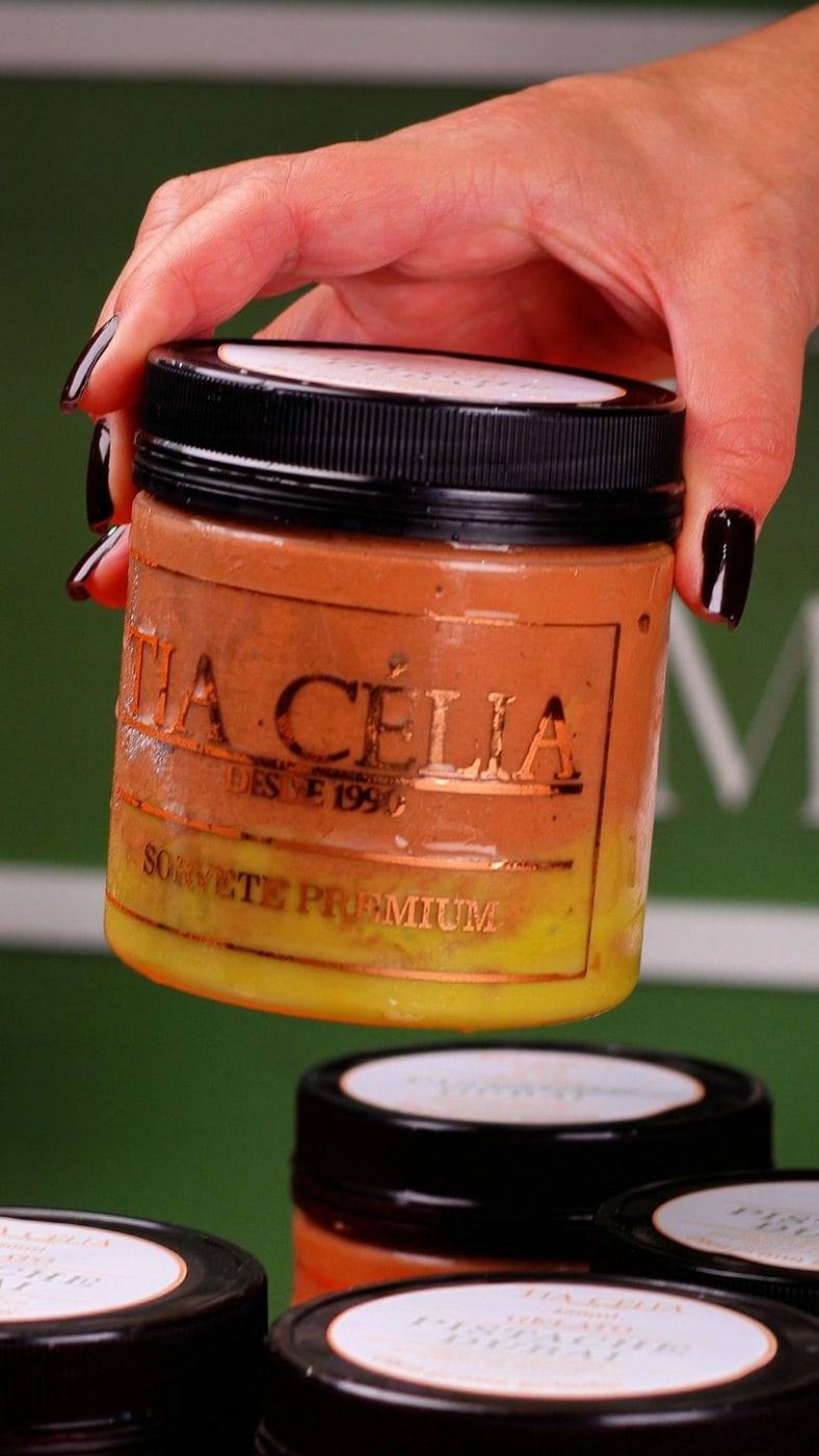 Mão segurando um pote de gelato Pistache Dubai da Tia Célia, com rótulo Sorvete Premium, sobre outros potes da linha.