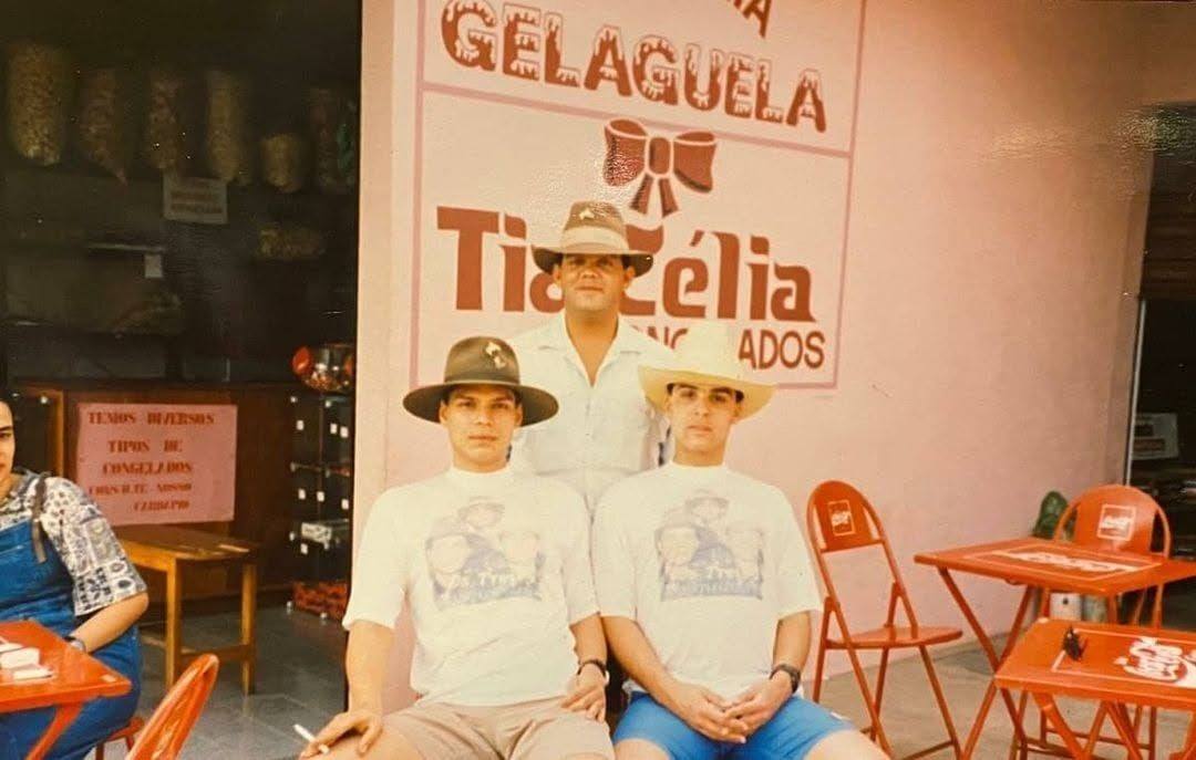 Clientes sentados diante da parede rosa com a placa da GelaGuela e o nome Tia Célia logo abaixo, ao lado de mesas e cadeiras laranja.