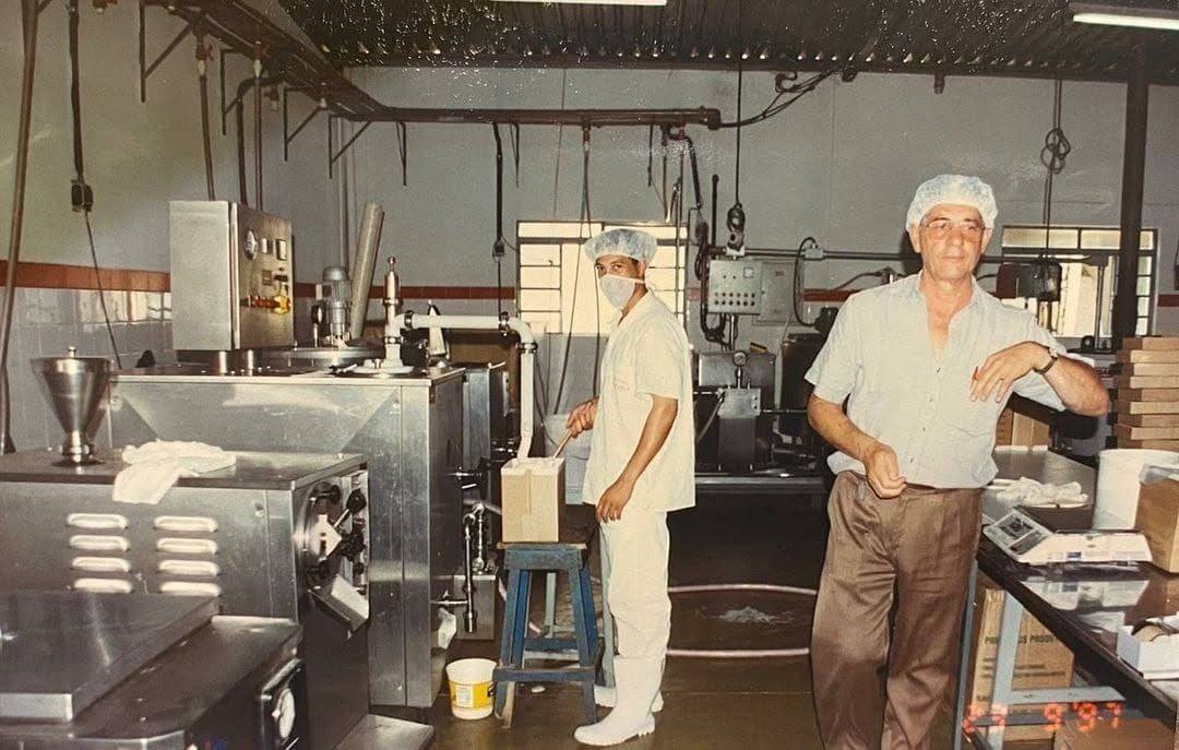 Tuban em pé na área de produção antiga, com máquinas de aço inox, ao lado de um funcionário de touca e uniforme branco.