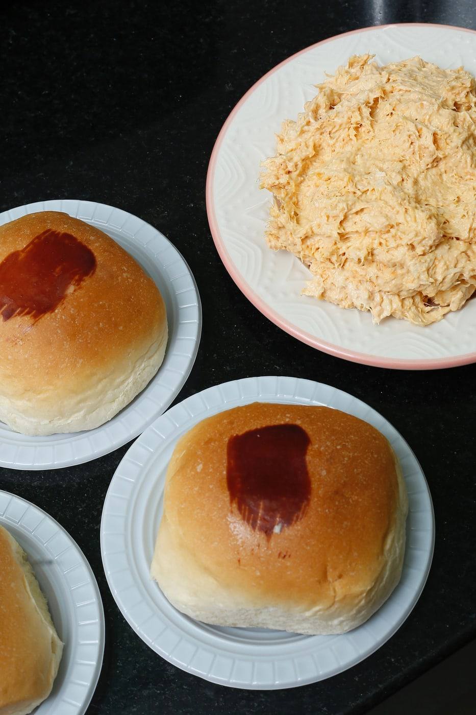 Pães de batata recheados com frango e requeijão em pratinhos brancos, ao lado de um prato de frango desfiado.
