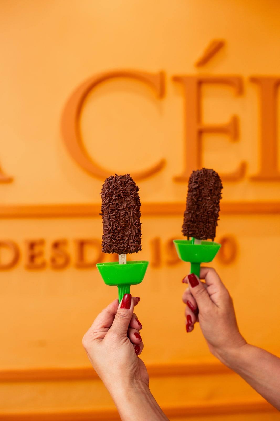 Duas mãos erguendo picolés de brigadeiro cobertos de granulado diante da parede laranja com o letreiro Tia Célia.