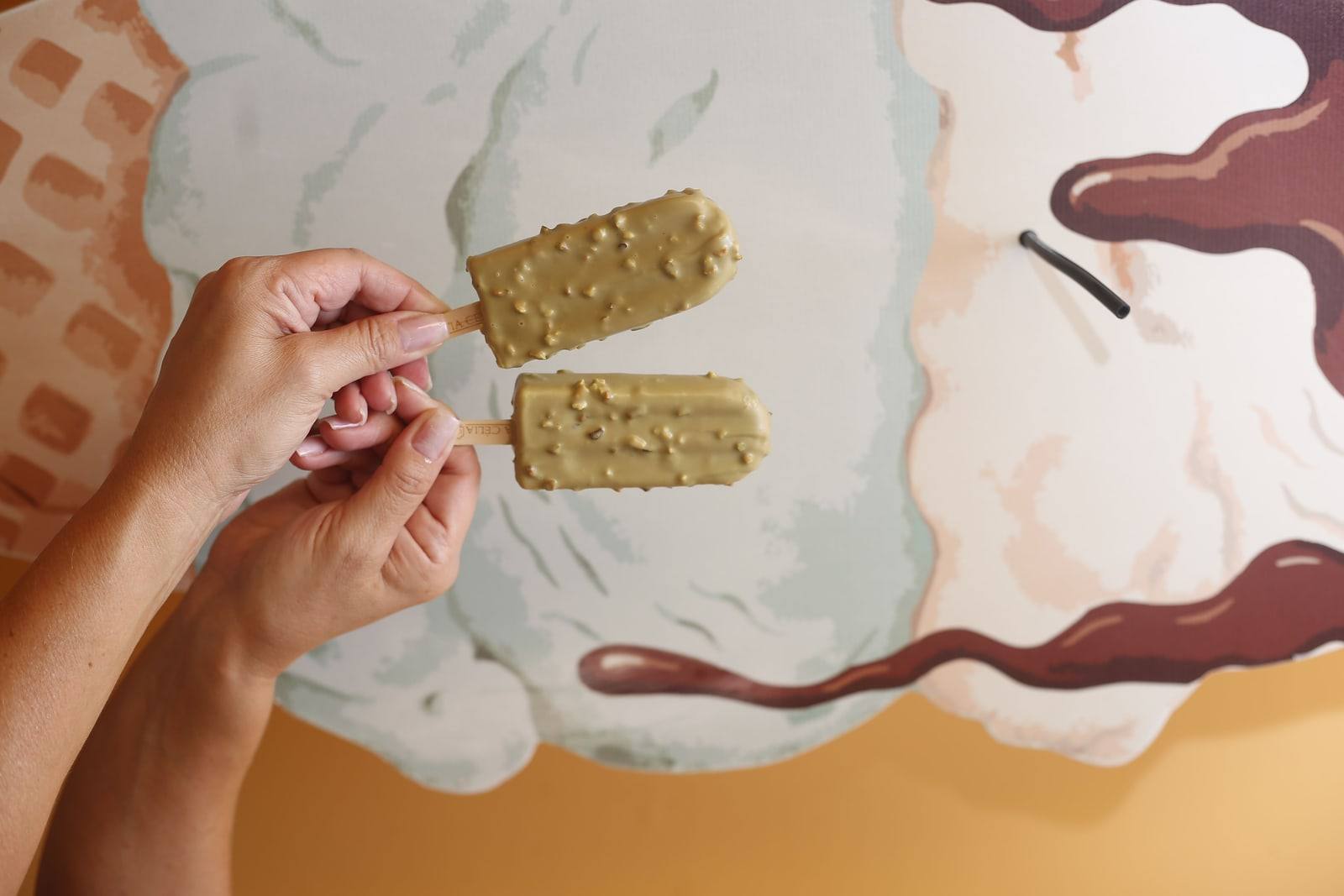 Mãos segurando dois picolés premium de pistache, cobertos e crocantes, diante de um painel ilustrado.