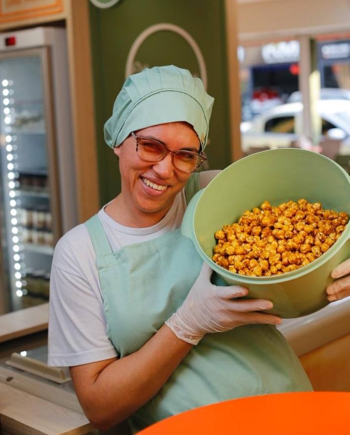 Nice, da produção da Tia Célia, sorridente de touca e avental verde-menta, segurando uma bacia de pipoca caramelizada.