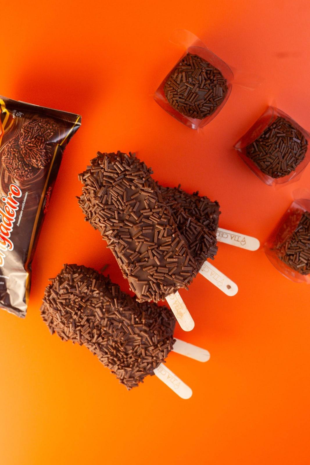 Picolés de brigadeiro cobertos de granulado de chocolate sobre fundo laranja.