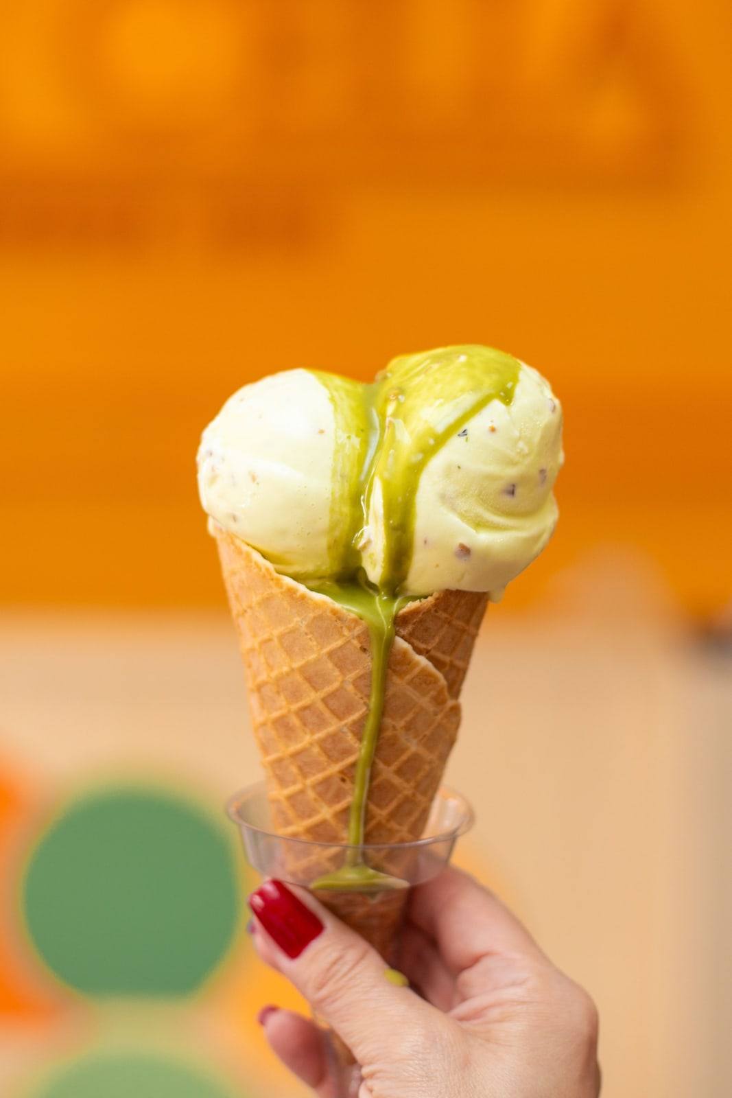 Cascão da Tia Célia com duas bolas de gelato de pistache, segurado diante da parede laranja da loja.